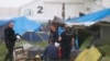 Greek policemen stand next to migrants at a makeshift refugee camp at the Greek-Macedonian border near the northern Greek village of Idomeni, May 24, 2016.